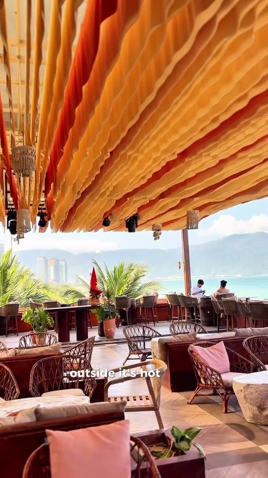 A DJ mixes music beside a laptop on a rooftop bar terrace overlooking the Da Nang coastline, with a Vietnamese flag and ocean view behind him.